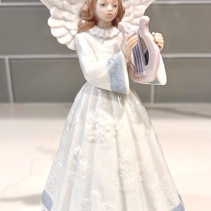 May include: A porcelain angel figurine with white wings and a long white dress with blue trim. The angel has brown hair and is holding a light purple harp. The dress has a floral pattern.