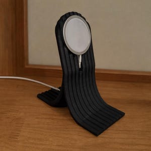 Pode incluir: Um suporte de carregamento preto e canelado com uma base de carregamento circular com borda prateada. O suporte está sobre uma superfície de madeira e um cabo de carregamento branco se estende da base. O suporte tem um design curvo único.