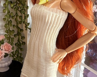 May include: A doll with long red hair wears a strapless, cream-colored, ribbed-texture mini dress. The doll is accessorized with a beaded necklace featuring yellow and silver beads with a central yellow pendant, and matching yellow star-shaped earrings.