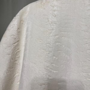 May include: A white faux fur garment featuring a dense, soft texture with a subtle, repeating embossed pattern throughout the fabric. The material appears thick and plush, with a consistent off-white color and a slightly shaggy, directional pile.