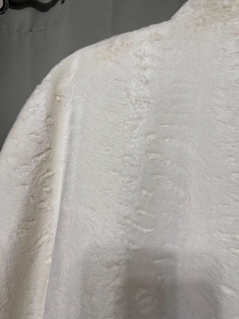 May include: A white faux fur garment featuring a dense, soft texture with a subtle, repeating embossed pattern throughout the fabric. The material appears thick and plush, with a consistent off-white color and a slightly shaggy, directional pile.