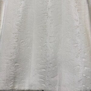 May include: A white faux fur jacket featuring a dense, soft texture with subtle vertical patterns throughout the material. The garment has a straight hemline and a smooth, satin-like lining visible along the bottom edge of the front panels.