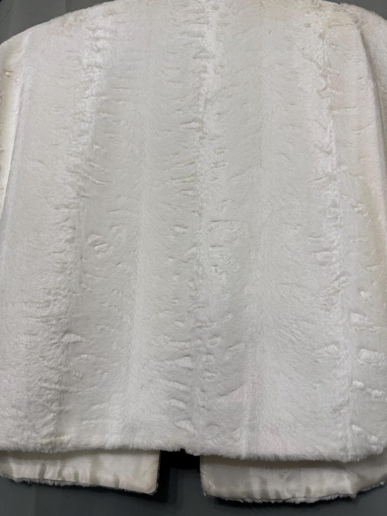 May include: A white faux fur jacket featuring a dense, soft texture with subtle vertical patterns throughout the material. The garment has a straight hemline and a smooth, satin-like lining visible along the bottom edge of the front panels.