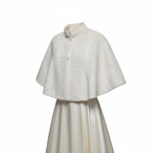 May include: A white cape with a textured surface and a high collar, featuring three round buttons down the front. The cape is worn over a smooth, floor-length white dress with a flared skirt, creating a layered appearance in this garment set.