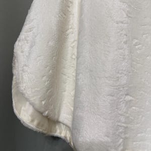 May include: A white faux fur capelet featuring a thick, plush texture and a smooth, satin-like fabric trim along the bottom edge. The garment has a soft, dense pile and a clean, solid white color throughout the entire piece, showing a draped silhouette.