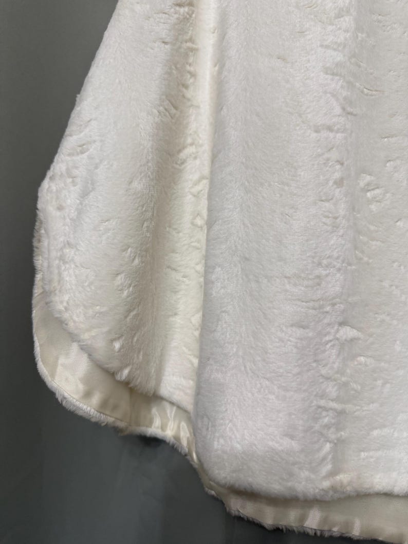 May include: A white faux fur capelet featuring a thick, plush texture and a smooth, satin-like fabric trim along the bottom edge. The garment has a soft, dense pile and a clean, solid white color throughout the entire piece, showing a draped silhouette.