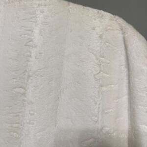 May include: A white faux fur coat featuring a dense, soft texture with subtle vertical patterns throughout the fabric. The garment is shown from the shoulder area, displaying the plush, thick pile of the material and its clean, bright white color.