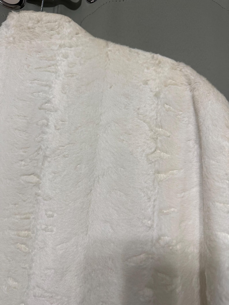 May include: A white faux fur coat featuring a dense, soft texture with subtle vertical patterns throughout the fabric. The garment is shown from the shoulder area, displaying the plush, thick pile of the material and its clean, bright white color.