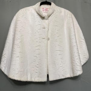 May include: A white cape featuring a textured, patterned surface and a high collar. The garment includes two round, fabric-covered buttons down the front. A label inside the neckline reads "Specialty House Fashion" in red script lettering.