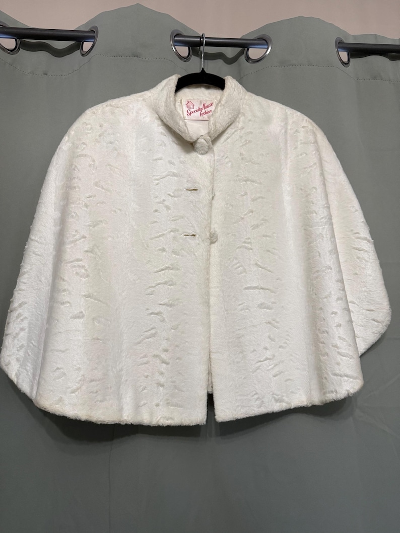 May include: A white cape featuring a textured, patterned surface and a high collar. The garment includes two round, fabric-covered buttons down the front. A label inside the neckline reads "Specialty House Fashion" in red script lettering.