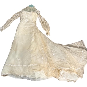 May include: A floor-length cream-colored gown featuring long sleeves, a high neckline with intricate lace detailing, and delicate floral embroidery scattered across the bodice and skirt. The dress has a flowing silhouette with a train.