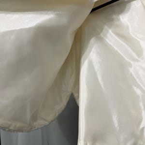 May include: A white faux fur bolero jacket featuring a smooth, shiny satin lining. The garment is displayed on a black hanger and includes a small white interior label with red text that reads Specialty Fashions near the neckline.