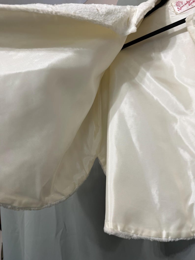 May include: A white faux fur bolero jacket featuring a smooth, shiny satin lining. The garment is displayed on a black hanger and includes a small white interior label with red text that reads Specialty Fashions near the neckline.