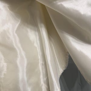 May include: A smooth, off-white satin fabric lining with a lustrous, reflective surface. The material is draped in soft, flowing folds, creating deep shadows and bright highlights across the surface, suspended from a thin black hanger bar.