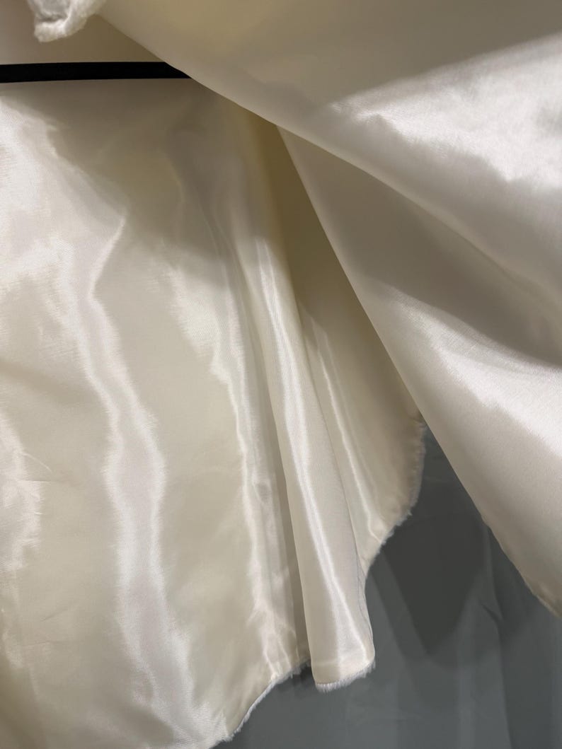 May include: A smooth, off-white satin fabric lining with a lustrous, reflective surface. The material is draped in soft, flowing folds, creating deep shadows and bright highlights across the surface, suspended from a thin black hanger bar.