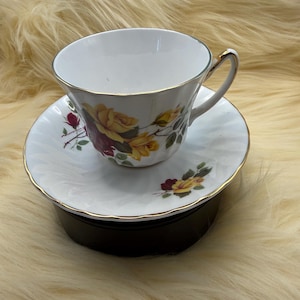 May include: White teacup and saucer set with a floral rose design in yellow and burgundy. The teacup and saucer have gold trim. The teacup is on a black stand, set against a fluffy, light-colored background.