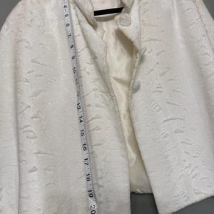May include: A white faux fur jacket with a textured surface, a stand-up collar, and two front buttons. A white measuring tape is placed vertically along the front, showing a length of approximately 22 inches from the collar to the bottom hem.
