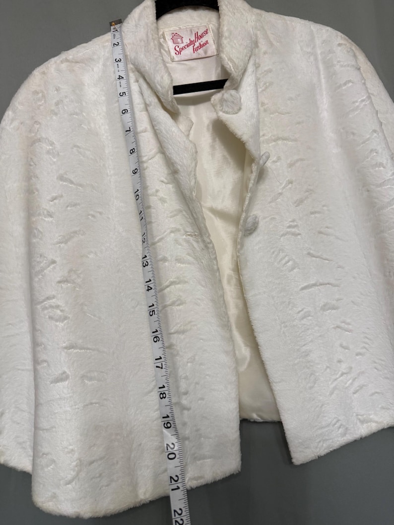 May include: A white faux fur jacket with a textured surface, a stand-up collar, and two front buttons. A white measuring tape is placed vertically along the front, showing a length of approximately 22 inches from the collar to the bottom hem.