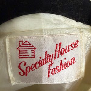 May include: A white garment label featuring a red embroidered graphic of a small house with a chimney. The text Specialty House Fashion is stitched in red thread across the center of the rectangular fabric tag, which is attached to a white garment.