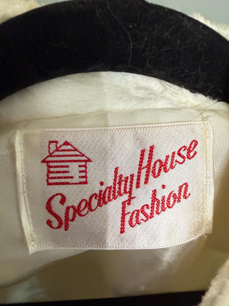 May include: A white garment label featuring a red embroidered graphic of a small house with a chimney. The text Specialty House Fashion is stitched in red thread across the center of the rectangular fabric tag, which is attached to a white garment.