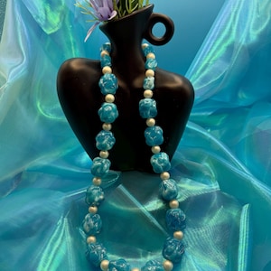 May include: A blue and white beaded necklace with a floral design. The necklace is draped in front of a black vase with purple flowers. The background is a sheer, turquoise fabric.