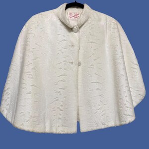May include: A white faux fur cape with a textured, wavy pattern throughout. The garment features a high collar, a single button closure at the neck, and a wide, flared silhouette. A label inside the collar reads Specialty House Fashion.