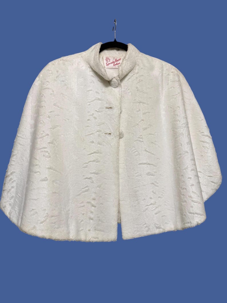 May include: A white faux fur cape with a textured, wavy pattern throughout. The garment features a high collar, a single button closure at the neck, and a wide, flared silhouette. A label inside the collar reads Specialty House Fashion.
