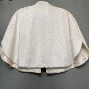 May include: A white faux fur capelet featuring a textured, shorn pile surface and a smooth, matching satin lining. The garment has a high collar, a rounded hemline, and an open front design, displayed on a hanger against a neutral backdrop.