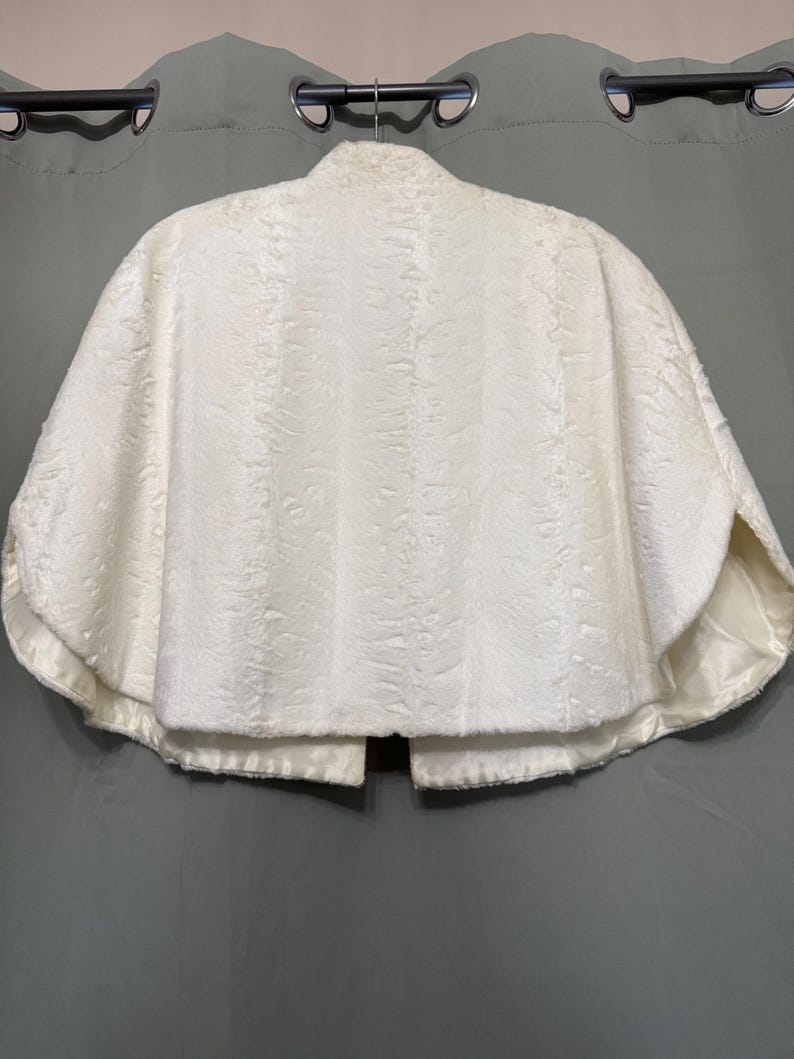 May include: A white faux fur capelet featuring a textured, shorn pile surface and a smooth, matching satin lining. The garment has a high collar, a rounded hemline, and an open front design, displayed on a hanger against a neutral backdrop.