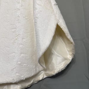 May include: A white faux fur garment featuring a soft, textured exterior surface and a smooth, satin-like lining visible at the side opening. The material has a dense, plush pile with a subtle directional pattern throughout the fabric.