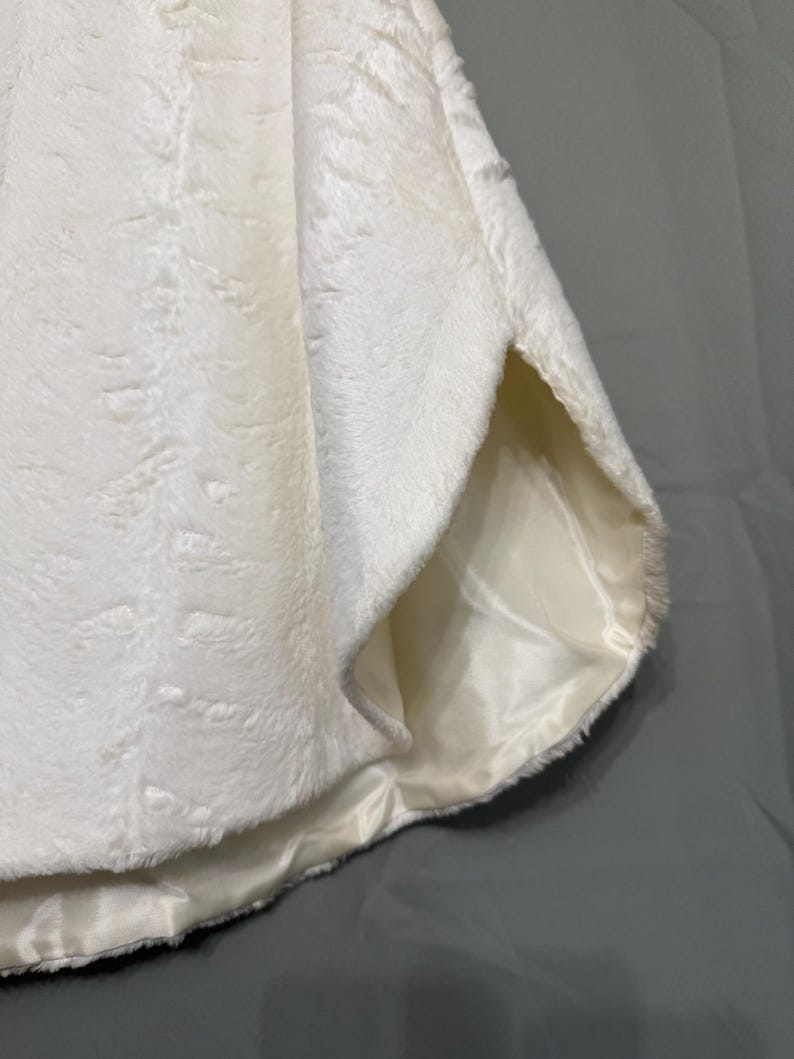 May include: A white faux fur garment featuring a soft, textured exterior surface and a smooth, satin-like lining visible at the side opening. The material has a dense, plush pile with a subtle directional pattern throughout the fabric.