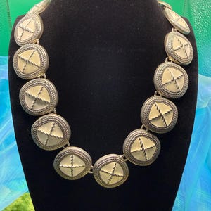 May include: A statement necklace featuring large, circular links. Each link has a cream-colored center with a cross design, framed by a silver-toned border and rope-like detailing. The necklace is displayed on a black mannequin.