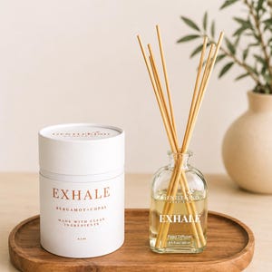 Könnte beinhalten: Eine weiße zylindrische Pappröhre mit der Aufschrift EXHALE BERGAMOT+COPAL 6.5 oz neben einem durchsichtigen Glas-Diffusor mit hellen Holzstäbchen, beschriftet mit EXHALE 6.5 fl oz/192 ml, auf einem ovalen Holztablett.