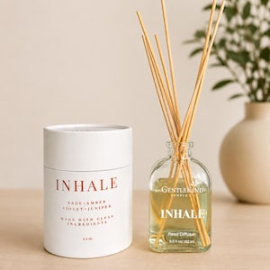 May include: A clear glass reed diffuser bottle filled with liquid and wooden reeds, accompanied by a white cylindrical cardboard tube. The label reads INHALE, SAGE + AMBER, VIOLET + JUNIPER, MADE WITH CLEAN INGREDIENTS, 6.5 oz/192 ml, GENTLEKIND CANDLE CO.