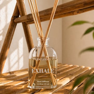 Puede incluir: Un difusor de varillas de vidrio transparente con la palabra "EXHALE" impresa en la parte delantera. La botella contiene un líquido de color claro y varias varillas de madera. La botella está etiquetada como "GENTLEKIND" y "Reed Diffuser 192 ml". La botella está sobre una estantería de bambú.