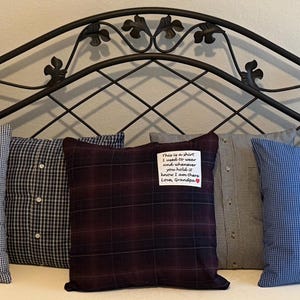 May include: Five decorative pillows made from repurposed shirts rest on a bed. The pillows feature various patterns, including gingham, plaid, and solid blue. One pillow has a handwritten note. The headboard is black metal with a decorative design.