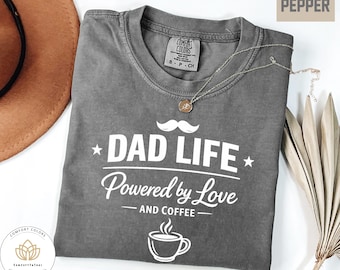 Könnte beinhalten: Ein graues Comfort Colors T-Shirt mit dem weißen Text "DAD LIFE Powered by Love AND COFFEE". Das Shirt hat einen Rundhalsausschnitt und kurze Ärmel. Eine goldene Halskette liegt über dem Shirt.