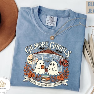 May include: A blue short-sleeve t-shirt featuring a graphic of two ghosts in a gazebo with the text "GILMORE GHOULS" and "WHERE YOU HAUNT, I WILL FOLLOW". The design includes autumn leaves, bats, a full moon, and a stack of books. The shirt has a crew neck.
