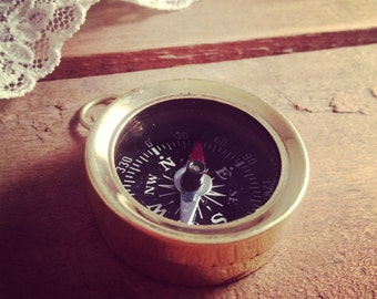 May include: A gold-toned compass with a black face and white markings. The compass has a red needle and is set against a wooden background.