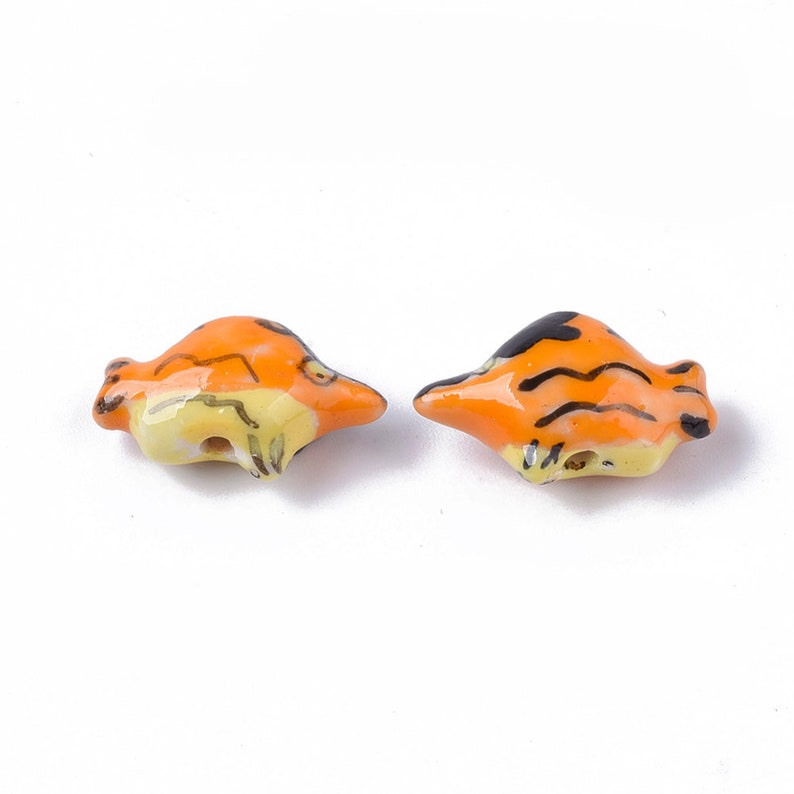 May include: Two ceramic beads shaped like fish with orange, yellow, and black tiger stripes. The beads have a hole through the center for stringing.