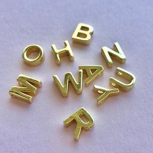May include: Gold letter beads in the alphabet letters B, H, M, N, O, R, W, A, Y, and U. The beads are scattered on a light pink surface.
