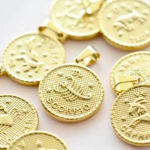 May include: A collection of round gold-toned metal pendants featuring embossed zodiac signs. Each pendant has a textured background, a raised astrological symbol, and the corresponding name printed below. One prominent pendant displays a scorpion and the text SCORPIO.