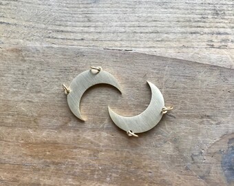 May include: Two gold crescent moon charms with jump rings. The charms are on a wooden surface.