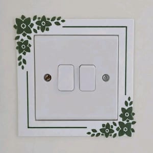 May include: A white double light switch cover plate featuring a dark green floral decal. The design includes a thin green border frame with clusters of stylized flowers and leaves positioned at the top-left and bottom-right corners of the plate.