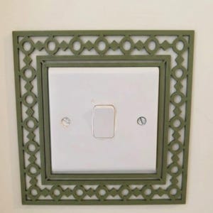 May include: A square olive green light switch cover plate featuring a decorative lattice pattern with repeating circles and diamonds. The plate surrounds a standard white rectangular light switch with two visible silver mounting screws.