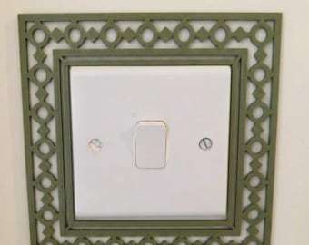May include: A square olive green light switch cover plate featuring a decorative lattice pattern with repeating circles and diamonds. The plate surrounds a standard white rectangular light switch with two visible silver mounting screws.