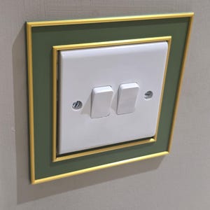 May include: A white double light switch mounted on a wall, framed by a square decorative border. The border features a dark olive green center panel with a thin, textured gold trim along the inner and outer edges, secured by two visible screws.