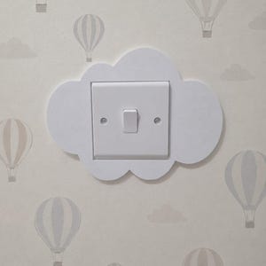 May include: A white light switch plate cover shaped like a cloud, mounted on a wall with light gray wallpaper featuring a pattern of hot air balloons and clouds. The switch plate is square with a single central toggle and two visible screws.