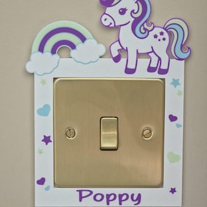 May include: A white light switch cover plate featuring a gold-colored metal switch. The plate is decorated with a purple and white unicorn, a rainbow with clouds, and small stars and hearts. The name Poppy is printed in purple at the bottom.