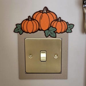Op de afbeelding: Een messingkleurige lichtschakelaarafdekking met een decoratieve bovenkant van drie oranje glinsterende pompoenen en groene bladeren. De vierkante plaat heeft een centrale tuimelschakelaar en twee zichtbare schroeven voor aan de muur.